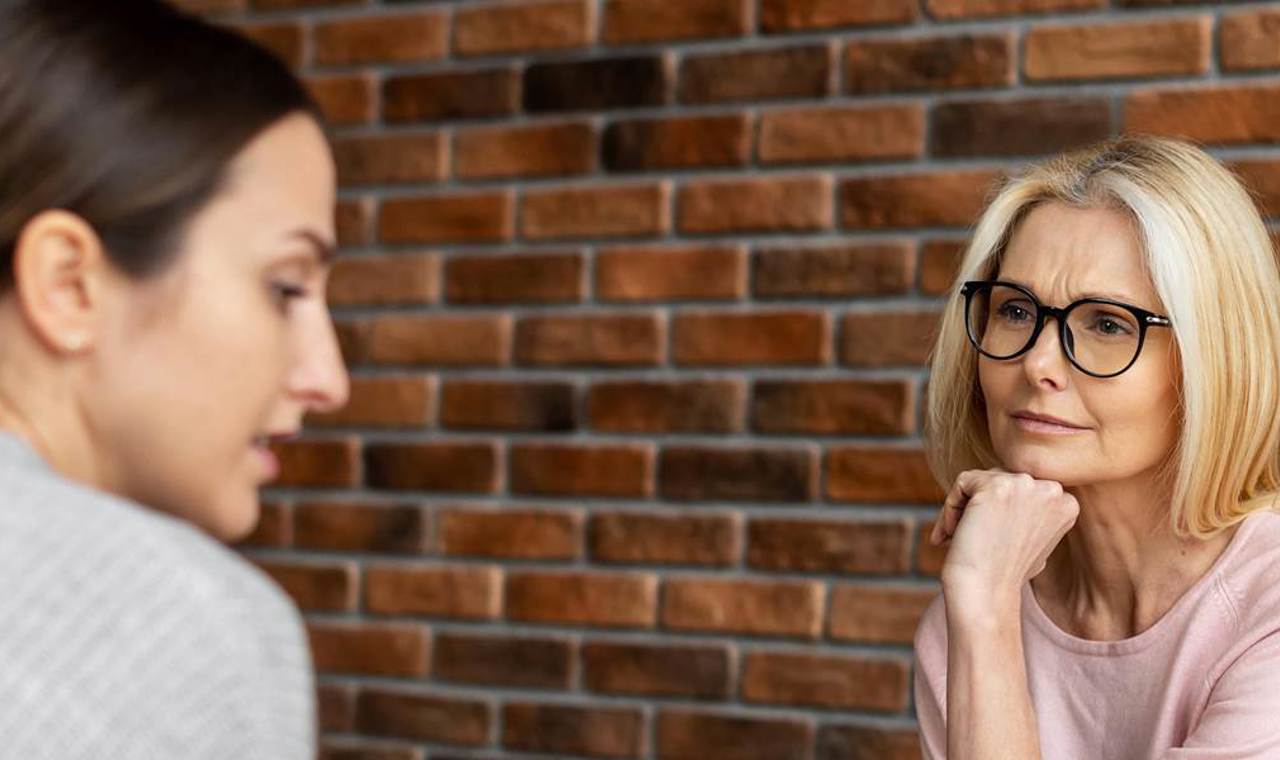 Twee vrouwen in gesprek, waarbij een oudere vrouw met bril aandachtig luistert naar de jongere vrouw, tegen een achtergrond van een bakstenen muur.